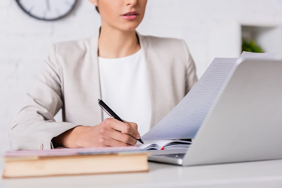 Cropped View Of Translator Writing In Notebook While Holding Document Near Laptop, Blurred Foregroun